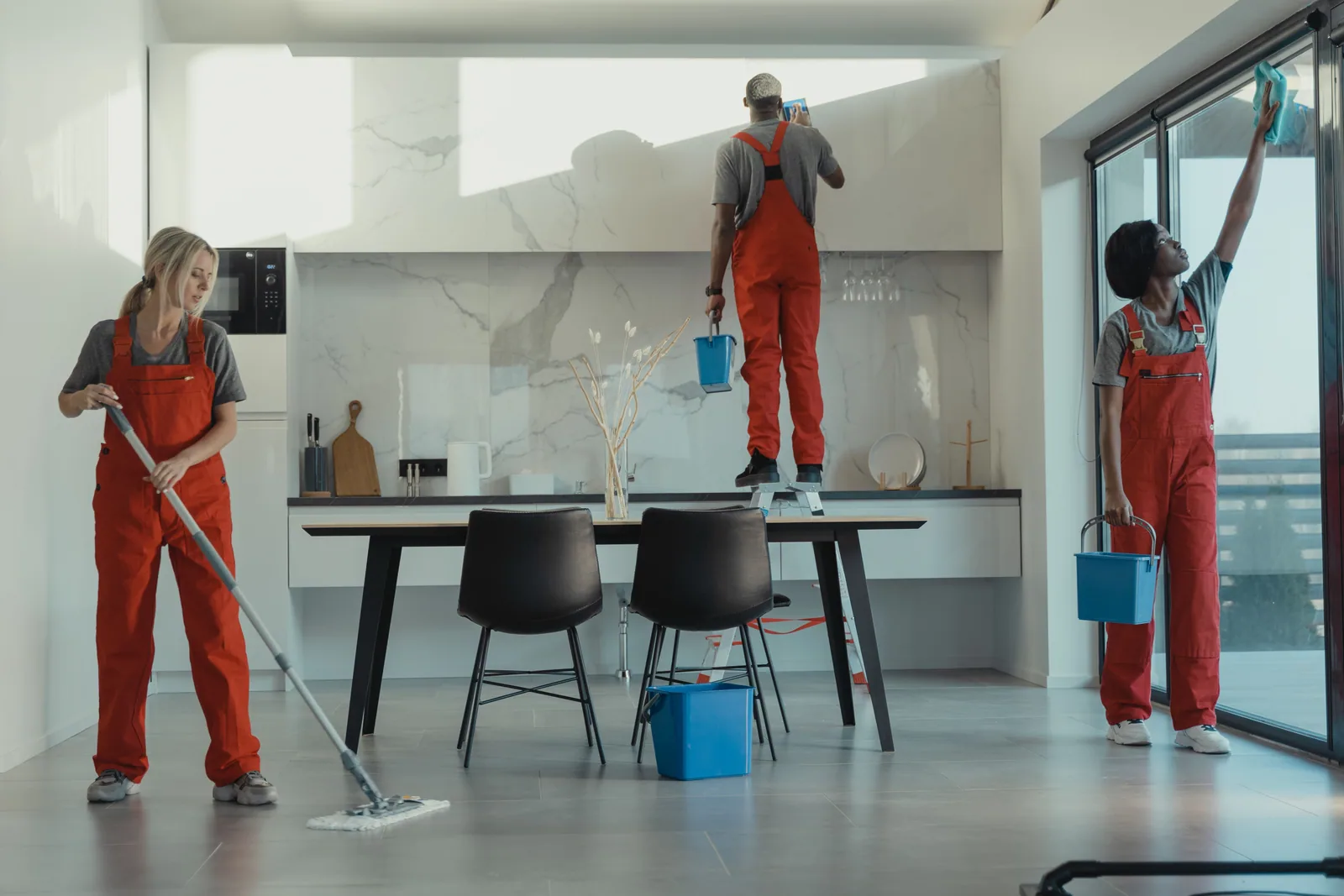 A three-person crew cleaning a kitchen: mopping, wiping the extractor and polishing the glass doors